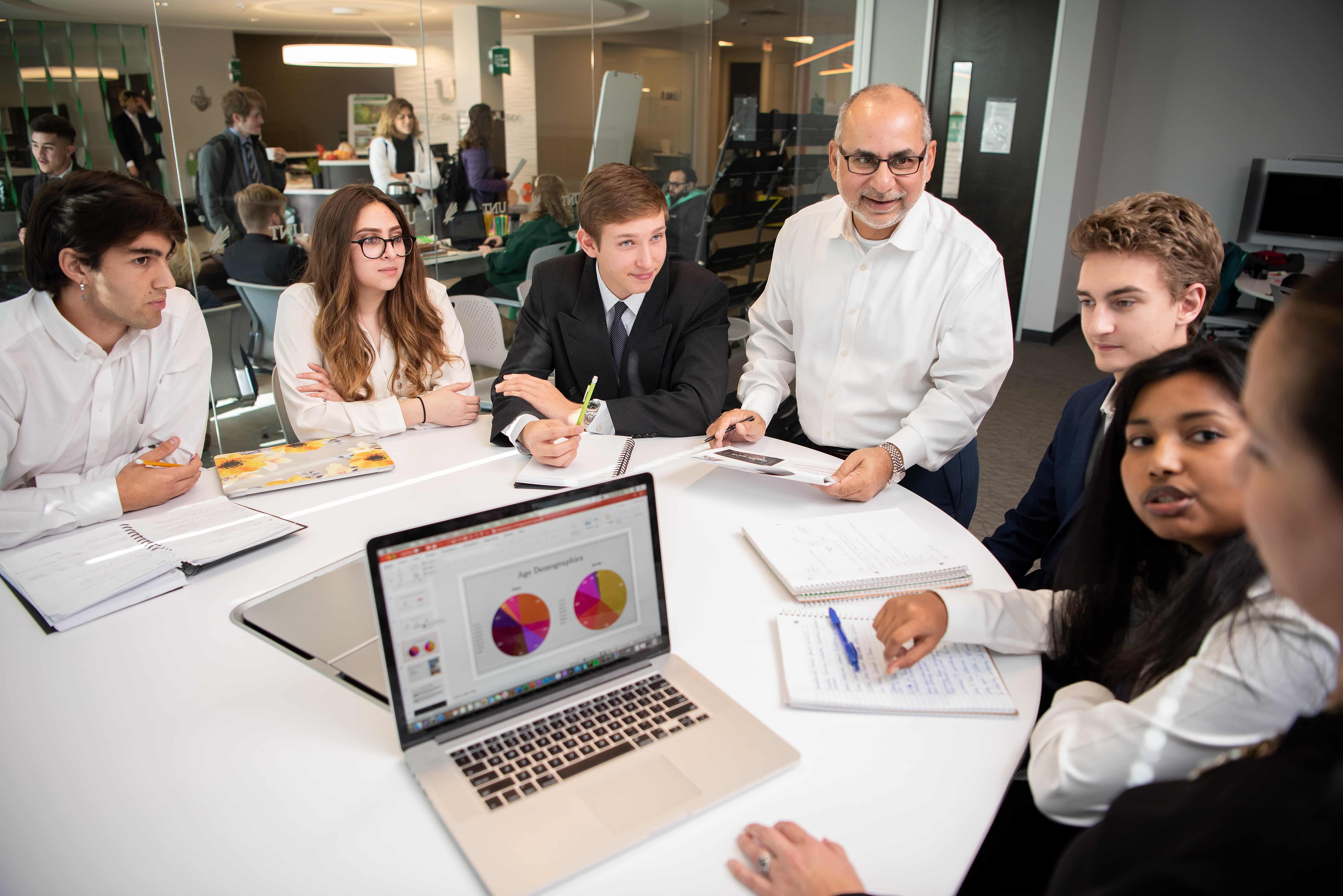 Students in business attire collaborating with a professor around a table, reviewing charts and notes on a laptop during a project-based learning session.