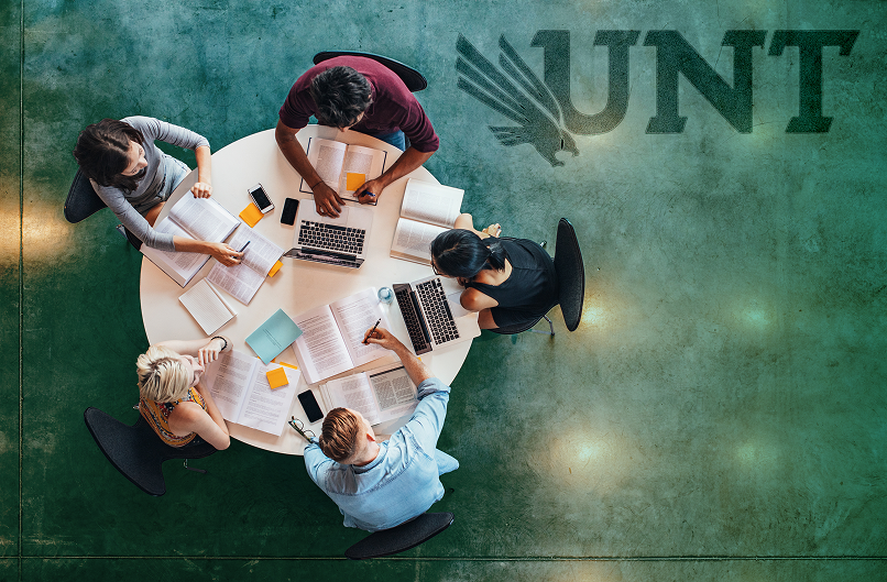 Five students sitting around a round table with laptops, books, and notes, studying together at UNT.